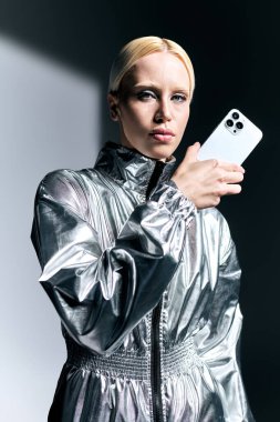 attractive extravagant woman in futuristic silver attire posing with phone and looking at camera clipart