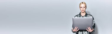 beautiful blonde female model in robotic silver outfit holding laptop and looking at camera, banner clipart