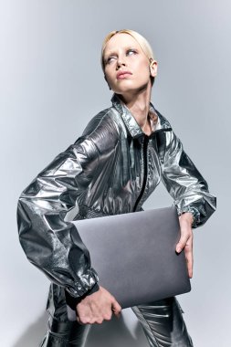 extravagant appealing woman in silver robotic clothing posing in motion with laptop and looking away clipart