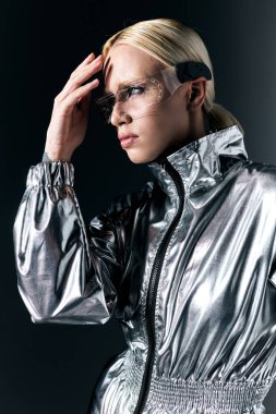 attractive blonde woman with sci fi glasses in futuristic attire looking away on dark gray backdrop clipart