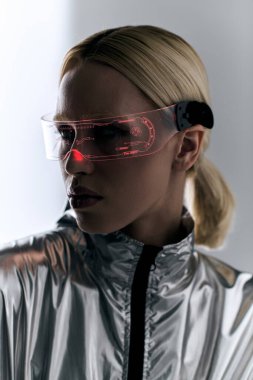 beautiful woman with futuristic glasses in robotic silver attire looking away on gray backdrop clipart