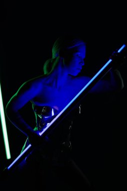 Gümüş renkli robot elbiseli güzel bir kadın mavi LED lamba çubuğuyla poz veriyor ve başka tarafa bakıyor.