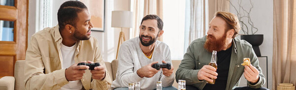 Three cheerful, interracial men sit around a table holding remotes, sharing laughs and camaraderie in a casual setting.