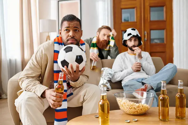 Three cheerful men of different races, casually dressed, discussing soccer tactics around a table with a soccer ball.