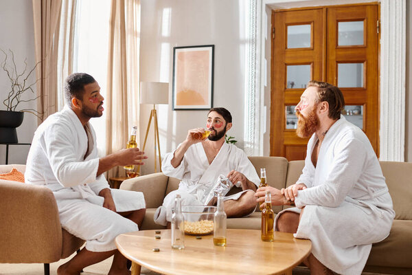 Three cheerful men of different backgrounds, in bathrobes, share laughter and camaraderie around a living room table.