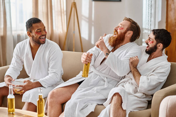 Three diverse, cheerful men in bathrobes sitting on top of a couch, enjoying a great time together.
