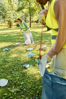 A socially active, diverse loving couple in safety vests and gloves clean a park as a group stands on a lush green field. clipart