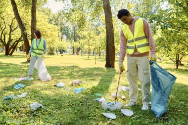 Güneşli bir günde parkı birlikte temizleyen güvenlik yelekleri ve eldivenleriyle sosyal olarak aktif, çeşitli bir çift..