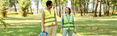 A socially active, diverse, loving couple in safety vests and gloves cleaning a park together, standing in the lush green grass. clipart