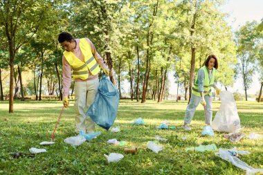 Socially active diverse couple wearing safety vests and gloves cleaning up trash in the park with a group of people. clipart