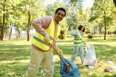 African american man in a yellow safety vest holds a blue bag while cleaning up in a park with his wife clipart