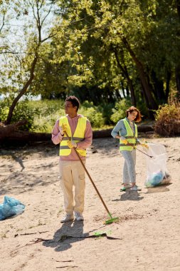 A couple in safety vests and gloves stands together in the sand, united in their commitment to cleaning the park. clipart