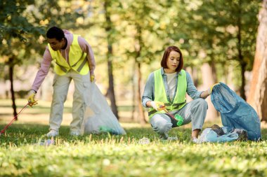 Diverse couple in safety vests and gloves stand on grassy field, actively participating in a park clean-up event. clipart