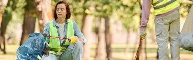 A woman, part of a socially active diverse loving couple, in a safety vest is picking up trash in a park. clipart
