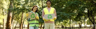 Güvenlik yelekleri ve eldivenleri olan bir çift, bir parkı temizlerken sosyal ve çevresel aktivizme olan tutkularını gösteren tabelalar taşıyorlar..