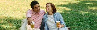 A serene moment captured as a diverse loving couple in vibrant attires sit together on the grass in the park. clipart