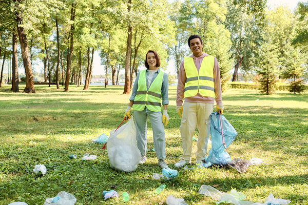 A socially active, loving couple in safety vests and gloves stands in the grass, cleaning the park together.