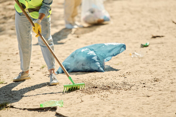 A woman in a safety vest and gloves sweeps up trash on the beach, embodying the spirit of environmental stewardship and care.