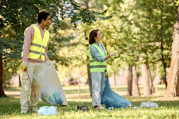 A loving, diverse couple in safety vests and gloves stand side by side on lush green grass, engaged in park cleanup.