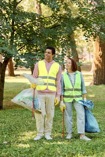 A socially active, diverse couple in safety vests and gloves stand in the grass, cleaning the park together in harmony.