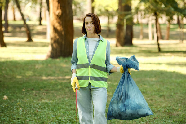 A woman in a green safety vest holds a blue bag while caring for nature in a park.
