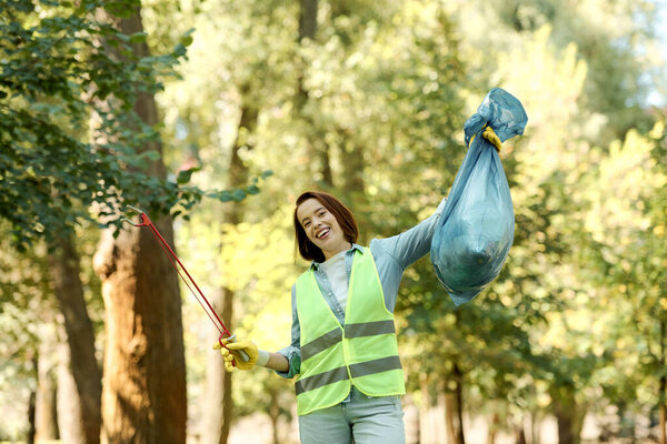 A woman in a bright safety vest is holding a vibrant blue bag while cleaning up a park with her partner in the background.