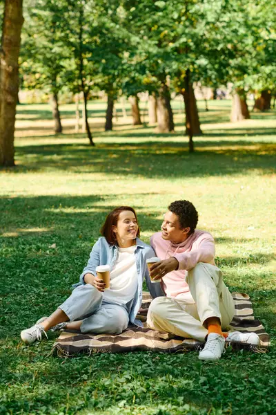 A diverse couple dressed in vibrant attire sitting on a blanket in the park, enjoying each others company.