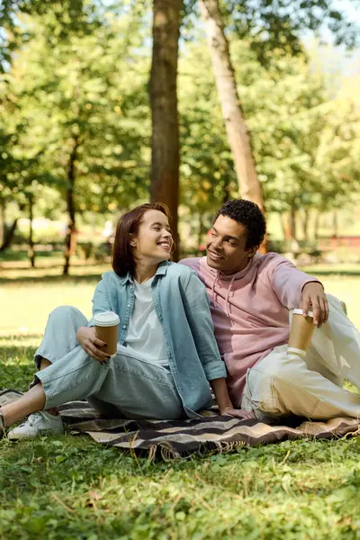 A diverse couple sits on a blanket in the park, enjoying each others company in vibrant attire.