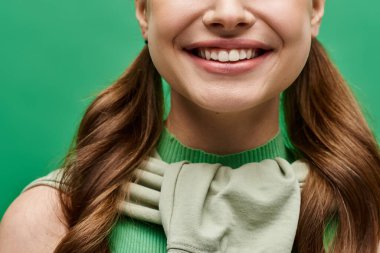 20 'li yaşlarda, uzun kahverengi saçlı genç bir kadın stüdyo ortamında yeşil bir gömlek içinde zarif bir şekilde poz veriyor..