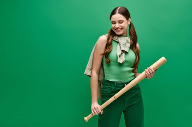 A young woman in her 20s stands holding a baseball bat in front of a green background. clipart