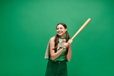 20 'li yaşlarda genç bir kadın beysbol sopasını canlı yeşil bir arka plana karşı tutarak güç ve kararlılık yayıyor..