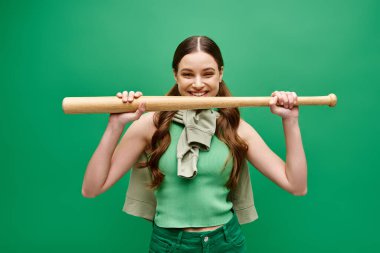 A young, beautiful woman in her 20s holds a baseball bat above her head in a dynamic pose against a green studio backdrop. clipart