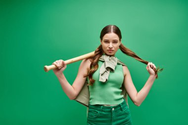 A young woman in her 20s confidently holds a baseball bat over her shoulder in a studio setting with green background. clipart