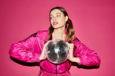 A stylish woman in her 20s wearing a pink jacket holding a glittering disco ball in front of a vibrant pink background. clipart