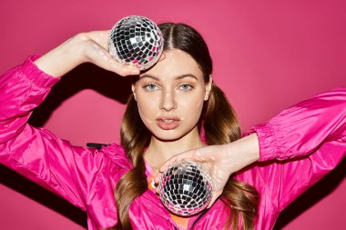 A young stylish woman in her 20s holding two disco balls in a studio with a pink background. clipart