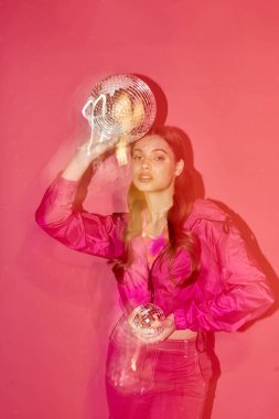 20 'li yaşlarda şık bir genç kadın, pembe bir gömlek giyiyor, pembe arka planda parlak gümüş bir top tutuyor..