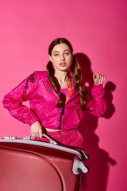 20 'li yaşlarda, şık bir şekilde pembe giyinmiş, elinde pembe arka planı olan bir bavul tutan bir genç kadın..