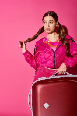 Pembe ceketli, 20 'li yaşlarda genç bir kadın. Stüdyo ortamında pembe arka planda kırmızı bavul tutuyor..
