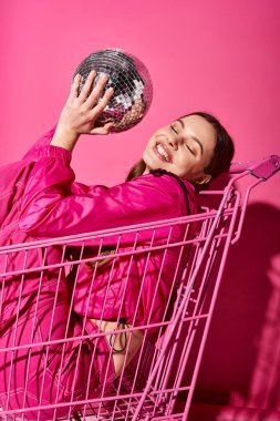 20 'li yaşlarda şık bir kız, pembe bir kıyafet giyiyor, pembe bir alışveriş arabasında canlı pembe bir arka plana karşı bir disko topu tutuyor..