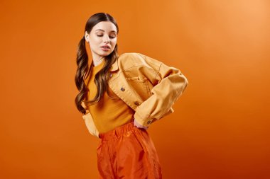 20 'li yaşlarda şık, sarı tişörtlü ve pantolonlu bir kadın turuncu arka planlı bir stüdyoda poz veriyor..