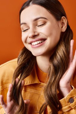 A young, stylish woman in her 20s smiles brightly while holding her hands together in a joyful expression, set against a vibrant orange background. clipart