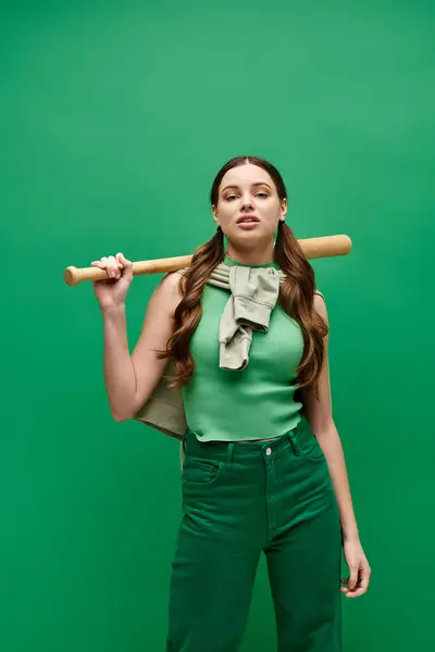 一位20多岁的年轻女子以自信的姿态将棒球棒扛在肩上 摆出一副绿色的画室姿势 — 图库照片