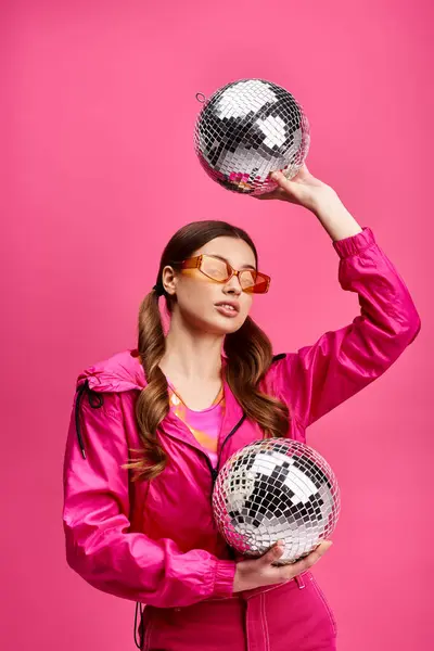 stock image Young woman in her 20s, stylish in a pink jacket, holds two disco balls against a vibrant pink background.