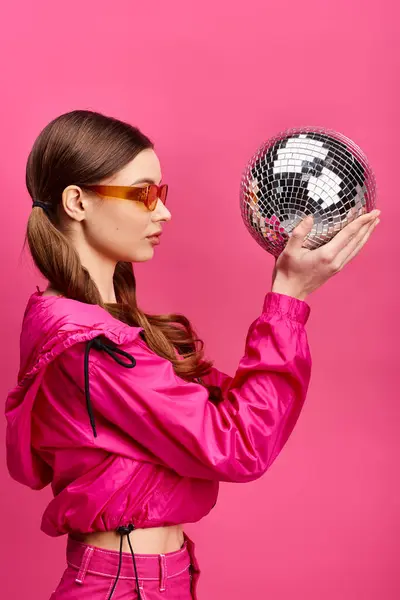 ピンクのジャケットを着た20代の若いスタイリッシュな女性は ピンクの背景を持つ活気に満ちたスタジオの設定でディスコボールを受け入れる — ストック写真