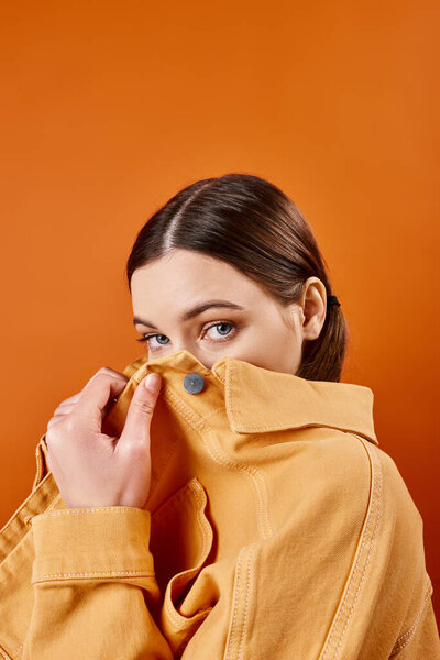 Young woman in her 20s, stylish in a yellow jacket, holds nose to nose. Studio setting with an orange background.