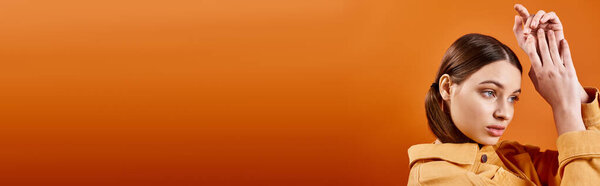 A young beautiful girl in her 20s in a yellow jacket enthusiastically raising her hands in a studio with an orange background.