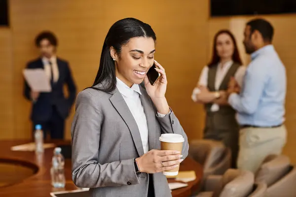 A woman in business attire talks on a cell phone while holding a cup of coffee.