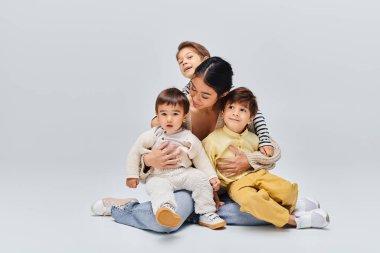 Genç bir Asyalı anne, çocukları ile birlikte gri arka planda bir stüdyoda yerde oturuyor..