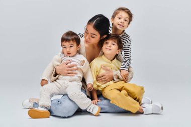 Genç bir Asyalı anne, çocuklarıyla birlikte gri bir zeminde, sakin bir stüdyoda oturuyor..
