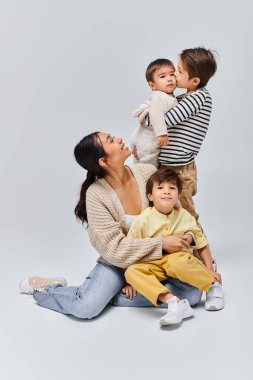 Genç bir Asyalı anne, çocuklarıyla birlikte gri bir arkaplanda, bir stüdyoda oturuyor..
